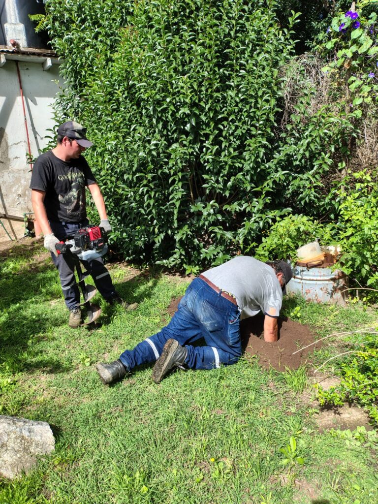 Dos operarios hacen pozo en patio de césped, uno sostiene la hoyadora y el otro saca los restos de tierra removida