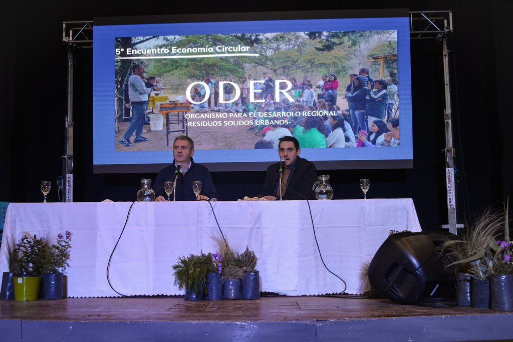 En el escenario del Encuentro el presidente del ODER y el coordinador del programa Involucrarnos, le hablan al público. De fondo hay una pantalla con la imagen de una charla en una escuela y el texto superpuesto que dice ODER; Organismo de Desarrollo Estratégico Regional, residuos sólidos urbanos.